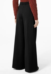 Persoon die hoge taille, wijde zwarte broek en een gestructureerde, langharige roestkleurige trui draagt, staande op een witte achtergrond.