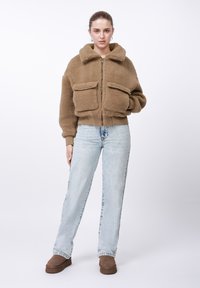 Nő barna, bolyhos cipzáras dzsekiben nagy zsebekkel, világoskék magas derekú farmernadrágban, barna belebújós cipőben, fehér háttér előtt áll.