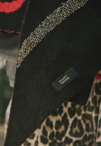 Juodas ir raudonas raštuotas audinys su audimo tekstūra, kuris turi etiketę "ROCKETT & GEORGE." Fone matyti leopardų rašto detalė.