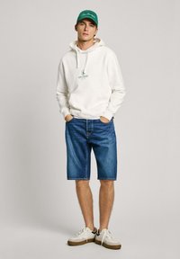 Sudadera blanca con capucha y logotipo verde, pantalones cortos de denim azul, y zapatillas blancas con acentos marrones, en un fondo neutro.