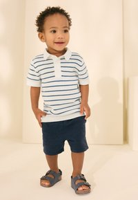 Ljusblårandig och vit randig pikétröja, marinblå shorts och blå sandaler med korksula på ett stående barn mot en neutral bakgrund.