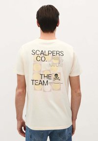 Krème-farvet bomuld t-shirt med sort og pastelfarvet grafisk tekst samt et kranium på ryggen, med et rektangulært mønster af cirkler.