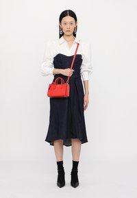 Abito blu navy con corpino aderente e gonna fluida, abbinato a una camicia bianca e una piccola borsa rossa. Tacchi a punta neri e calzini completano il look.