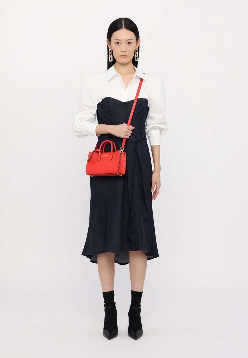 Abito blu navy con corpino aderente e gonna fluida, abbinato a una camicia bianca e una piccola borsa rossa. Tacchi a punta neri e calzini completano il look.