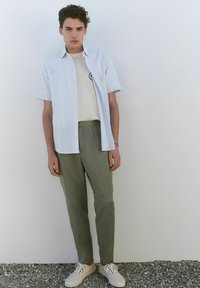 Chemise à manches courtes, rayée bleu clair, sur un t-shirt beige, associée à un pantalon vert olive et des baskets blanches. Design simple et moderne.