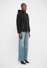 Veste en duvet noire avec capuche, ornée d'un design en chevrons et de détails de fermeture éclair dorés. Associée à un jean large bleu clair et des bottes noires à bout pointu.
