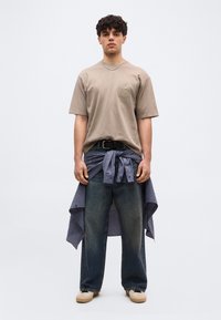 Νεαρός άνδρας που φοράει μια φαρδιά μπεζ μπλούζα, σκούρα μπλε τζιν, μπεζ αθλητικά παπούτσια και ένα μπλε καρό πουκάμισο δεμένο στη μέση του.