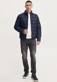 Marineblå puffjakke med glidelås, horisontal søm, og høy krage, båret over en hvit skjorte og kombinert med grå jeans og svarte sneakers.