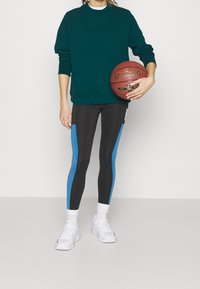 Donkergroen sweatshirt met lange mouwen, gecombineerd met zwarte leggings met blauwe en rode zijpanelen en witte sportschoenen. De persoon houdt een basketbal vast.
