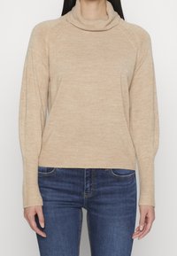 Beżowy sweter dziany z dekoltem w kształcie stójki, z prążkowanymi mankietami i dolnym wykończeniem, o luźnym kroju i lekko teksturowanej tkaninie. W połączeniu z ciemnoniebieskimi dżinsami.