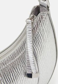 Srebrna torebka o metalicznym, fakturowanym wykończeniu, zamykana na zamek błyskawiczny, z płaskim paskiem ozdobionym kwadratowym akcentem. Kompaktowy, półokrągły kształt.