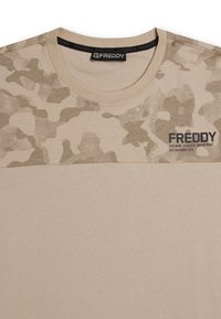 Camiseta beige con patrón de camuflaje en la parte superior, cuello redondo y mangas cortas. Logo y texto "Freddy" en negro en el frente. Textura suave.