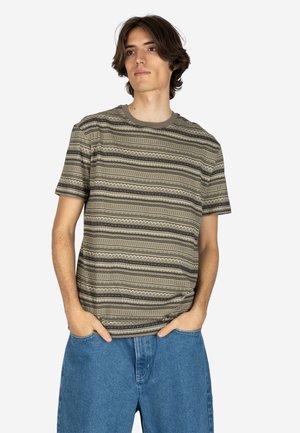 Junger Mann mit schulterlangem Haar, der ein grünes, gemustertes Kurzarmhemd und blaue Jeans trägt, die Hände in den Taschen, blickt leicht nach rechts.
