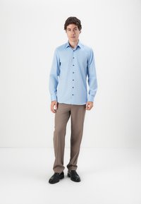 Hellblaues Hemd mit langen Ärmeln und dunklen Knöpfen, kombiniert mit beigen Anzughosen und schwarzen Loafers, vor einem weißen Hintergrund stehend.