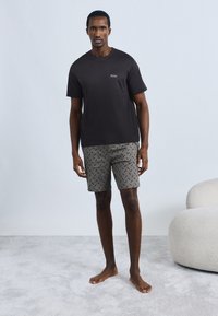 Μαύρο βαμβακερό T-shirt με μικρό λογότυπο στο στήθος, συνδυασμένο με γκρι σορτς με σχέδιο. Το μοντέλο στέκεται ξυπόλητο σε ανοιχτόχρωμη επιφάνεια.