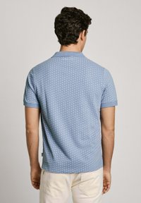 Světle modré polo tričko s opakujícím se malým bílým vzorem, krátké rukávy a límec. Hladká textilie.