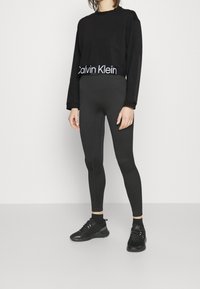 Svart croppad sweatshirt med en vit Calvin Klein-logotyp, kombinerad med svarta leggings och svarta träningsskor. Slät tyg med en figurnära design.