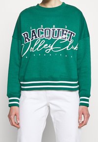 Sudadera de algodón verde con gráfico de "Racquet Volley Club" en azul marino y blanco, con puños y dobladillo acanalados, y acentos a rayas.