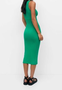 Femme dans une robe midi verte ajustée sans manches, portant des sandales plates noires à lanières, se tenant devant un fond blanc, vue de dos.