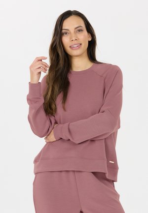 Jonge vrouw met lang bruin haar, een mauve sweatshirt en bijpassende broek dragend, glimlachend met één arm gekruist en de andere omhoog bij haar gezicht.