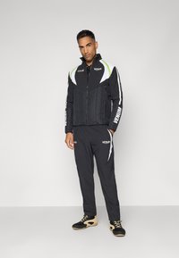 Black athletic tracksuit with a quilted jacket featuring white and neon green accents, paired with matching pants and black, textured shoes.