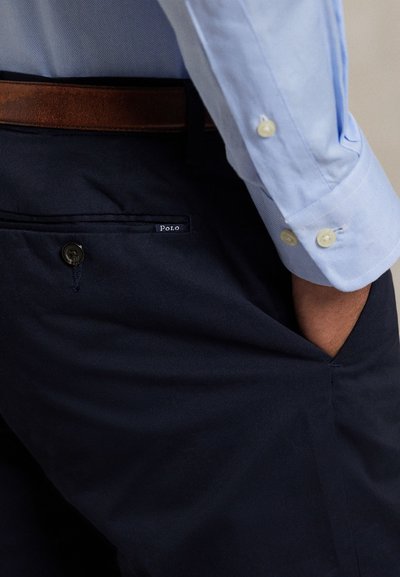 Muž v tmavě modrých kalhotách s náplastí "Polo" na zadní kapse, hnědým páskem a světle modré košili s dlouhými rukávy a zapnutými manžetami, rukou v kapse.