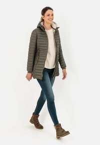 Gewatteerde olijfgroene jas met een ritssluiting aan de voorkant, capuchon en gestikte design. Gecombineerd met blauwe jeans en bruine wandelschoenen. Neutraal blouse eronder.