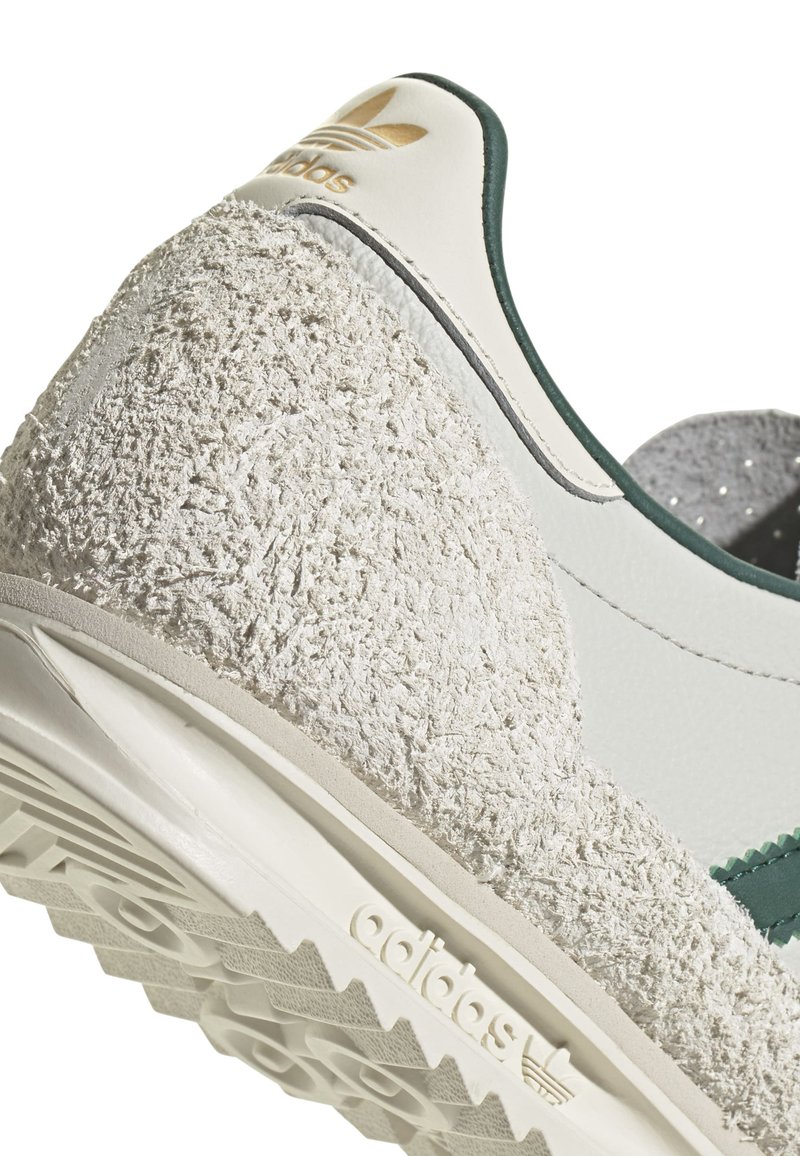 Baskets blanches avec un talon en suède gris texturé, des accents verts et une tige lisse. Présente une semelle en caoutchouc à motif et un logo en relief.