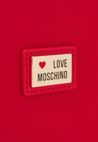 Czerwona skórzana torba z akcentem w postaci złotej prostokątnej zawieszki, na której widnieje tekst "LOVE MOSCHINO" oraz mała grafika serduszka nad napisem.