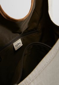 Indvendig udsigt af en brun Kipling taske med lynlåslommer og stofmærke med Kipling Live.Light branding.