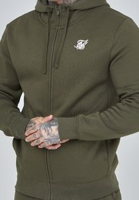 Sudadera con cremallera de color verde oliva, hecha de tela suave, que cuenta con un bolsillo frontal, capucha con cordón y un pequeño logo blanco en el pecho.