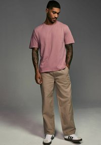 Tricou din bumbac roz, croi lejer, purtat cu pantaloni bej și teniși albi cu dungi negre. Mâneci scurte, gât rotund, textură netedă.