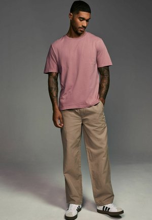 Lyserød bomulds T-shirt, løs pasform, båret med beige bukser og hvide sneakers med sorte striber. Korte ærmer, rund hals, glat tekstur.