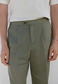 Pantalons vert olive avec une texture lisse, dotés de plis, d'un zip à l'avant et d'une fermeture à bouton décoratif.