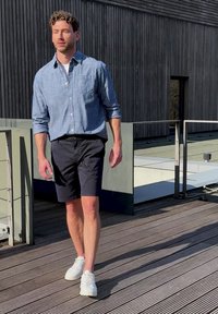 Homme marchant sur une terrasse en bois, portant une chemise bleue à manches longues, un short noir et des baskets blanches près d'un bâtiment moderne avec des panneaux verticaux foncés.
