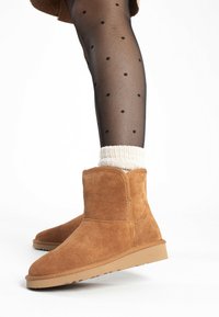 Botas de ante marrón hasta la pantorrilla con puntera redonda y suela de goma plana, combinadas con medias transparentes negras con lunares y un calcetín blanco de canalé.