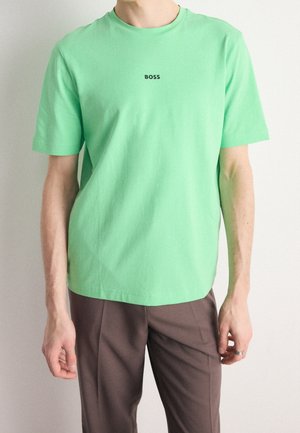 Mann trägt ein hellgrünes T-Shirt mit dem Wort "BOSS" in schwarzer Schrift in der Mitte und braune Hose vor einem einfarbigen weißen Hintergrund.
