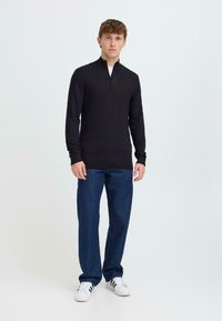 Pull en tricot noir avec une fermeture éclair quart, dotée d'un col haut et de poignets côtelés, associé à un jean droit bleu marine et des baskets blanches.