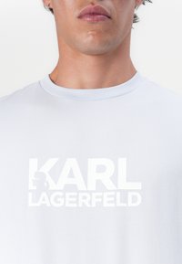 Šviesiai mėlyna marškinėlių medžiaga, pagaminta iš lygaus audinio, su dideliu baltu logotipu, kuriame parašyta "KARL LAGERFELD" ir silueto grafika.