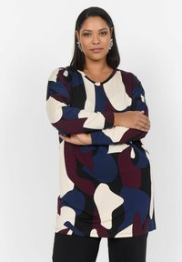 Tunique à manches longues dans un motif abstrait crème, marine et bordeaux, avec un décolleté en V et un tissu doux. Elle est portée avec un pantalon noir en dessous.