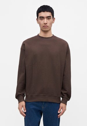 Jeune homme aux cheveux noirs courts portant un sweat-shirt marron uni à manches longues et un jean bleu, debout devant un fond blanc.
