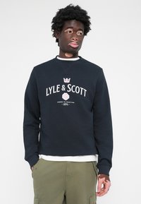 Navy sweatshirt from Lyle & Scott with white logo and crown graphic. Ribbed cuffs, round neckline, and soft texture. Layered over a white shirt.