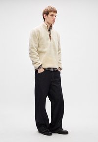 Creme fleece pullover med halv-lynlås og brun lynlås, lagd over en ternet skjorte. Stylet med løse mørke denimjeans.