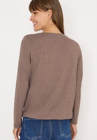 Maglietta marrone a maniche lunghe con collo a giro, realizzata in tessuto morbido. Il retro presenta un orlo leggermente curvo, abbinata a jeans blu.