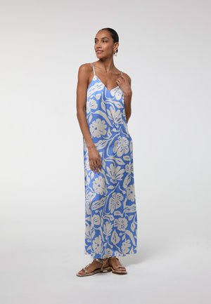 Mujer con un vestido largo azul con estampado floral blanco y tirantes finos, de pie con una mano cerca de su clavícula, llevando sandalias.