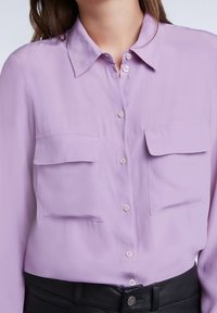 Camicia button-up lavanda con colletto, dotata di due tasche sul petto e una texture liscia. Presenta una chiusura frontale con bottoni e un design aderente.