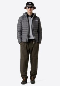 Grå puffjacka, grå sweatshirt, mörkgröna byxor, svart bucket hat och svarta solglasögon. Strukturerade material och avslappnad passform.
