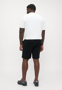 Svarte shorts med en glatt tekstur, kombinert med en hvit T-skjorte med korte ermer, iført svarte joggesko. Bakfra, nøytral bakgrunn.