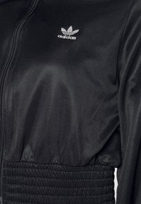 Svart Adidas träningsjacka med dragkedja och elastiskt ribbat midjeband, med vit Adidas trefoil-logotyp på bröstet.