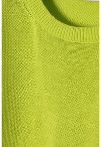 Detailní záběr na měkkou pletenou látku v jasně limetkově zelené barvě se vroubkovaným kulatým výstřihem, zdůrazňující texturu a detaily švů.